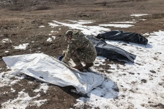 Чоловік у камуфляжі на засніженій ділянці землі оглядає тіло, загорнуте в білий мішок, поряд лежать ще два чорні мішки з тілами.