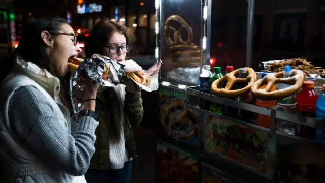 Două fete mâncând crenvurști în fața unui chioșc de fast food.