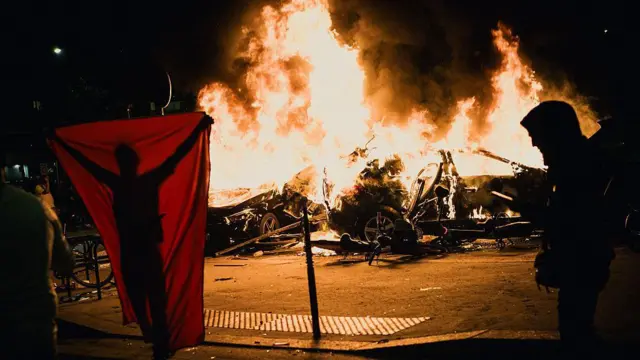 Un coche arde en llamas mientras dos hombres pasan por delante.