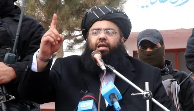 تصویر مولوی عبدالکبیر حین سخنرانی. او ریش دراز سیاه دارد و دستار سیاه به سر کرده است. او در پشت مایکروفونها ایستاده و انگشت دستش را به علامت هشدار بالا برده است.