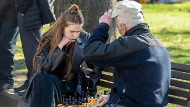 Молода жінка і старший чоловік сидять на лавці у парку, грають у шахи. 