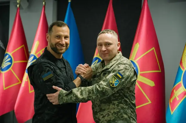 Двоє чоловіків у військовій формі стоять у приміщенні та тиснуть одне одному руки, охопивши їх обома долонями. Один одягнений у темну форму, інший — у піксельний камуфляж із нашивкою прапора України на рукаві. На задньому плані розташовано кілька червоних прапорів із військовою символікою та один синій прапор.