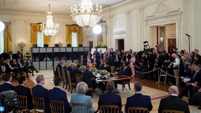 複数のシャンデリアが天井から下がり、華やかな装飾の施されたホワイトハウスの広間の、中央に長い木製のテーブルが置かれ、トランプ氏の前にゼレンスキー氏が座り、その周りに欧州首脳らが座っている。このテーブルの周りを、木製の椅子に座った各国政府のスタッフが囲み、写真の奥には通訳ブースがある。入口の周辺には、ロープの後ろに報道陣が集まり、カメラやマイクを構えている。
