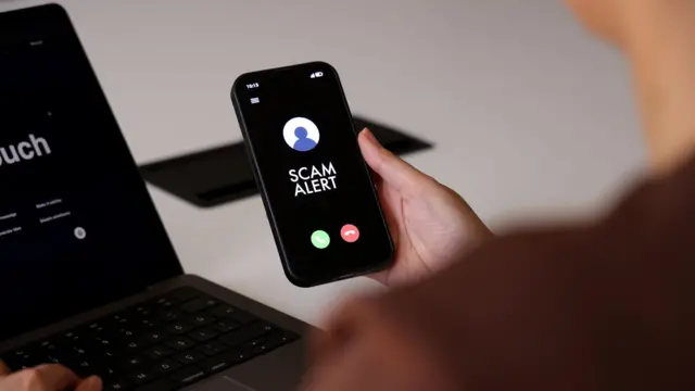 Bir kişi, gelen arama ekranında "Dolandırıcılık Uyarısı" mesajı gösteren bir akıllı telefon tutuyor.