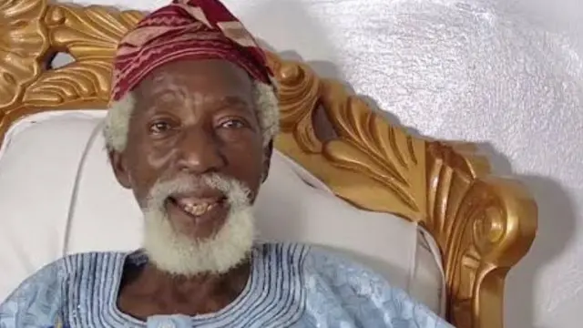 Bàbá Gébú joko sori aga, ó wọ aṣọ leesi aláwọ̀ ojú ọrùn, ó sì dé fìlà aṣọ oke aláwọ̀ waini