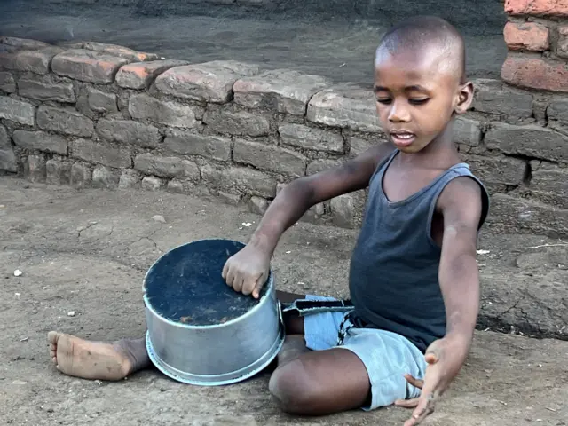A young boy sits on the ground in shorts and a vest playing with a cooking pot. 