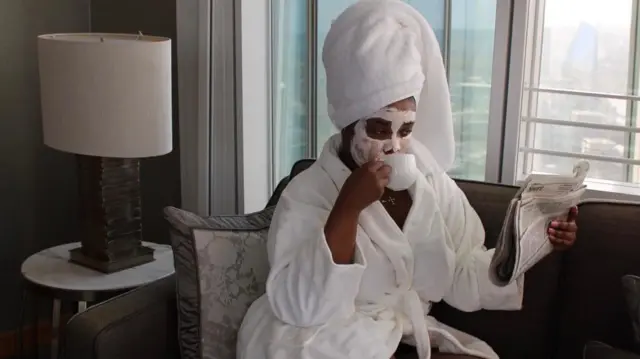 A woman is sat on a sofa in a white bath robe, having a cup of tea whilst wearing a face mask and reading the newspaper. 