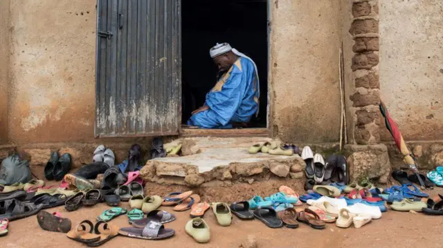 Tarin takalma a ƙofar wani masallacin da aka gina da ƙasa, ga wani mutum can bakin ƙofa daga ciki yana zaman tahiya