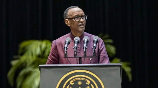 Perezida w'u Rwanda Paul Kagame ageza ijambo ku badipolomate, i Kigali, ku wa kane, ku itariki ya 16 Mutarama (1) mu mwaka wa 2025. Yambaye ishati y'iroza n'indorerwamo, ahagaze imbere y'ameza maremare ariho indangururamajwi eshatu, inyuma ye haraboneka ururabo rw'amababi y'icyatsi kibisi, n'umwijima wa nijoro. 