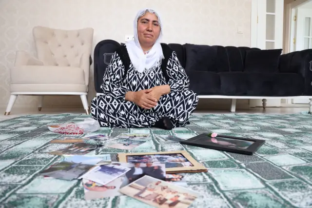 Emine Al, oğlunun fotoğraflarını gösteriyor. Yerde oturmuş, önünde fotoğraflar var.