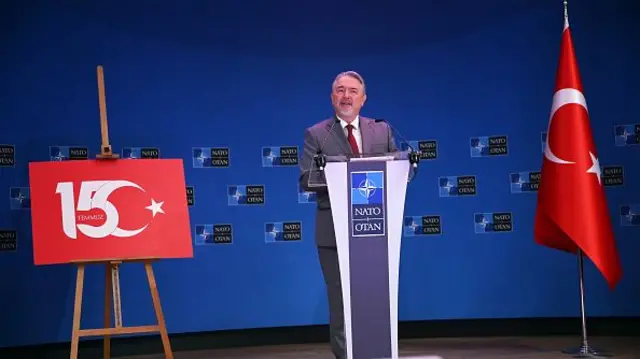 Basat Öztürk takım elbiseli ve NATO yazan bir kürsüde 15 Temmuz darbe girişimi anmasında konuşuyor arka planda NATO logoları var, sağ yanında Türk bayrağı sol yanında 15 ve ay yıldızın birleştiği bir tablo var. 15 temmuz 2025. 