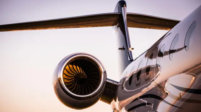 A detail in close-up of the side of a luxury jet at what looks like sunrise or sunset - the image is focused on one of the engines of the plane, the body of which is reflecting the scene around it