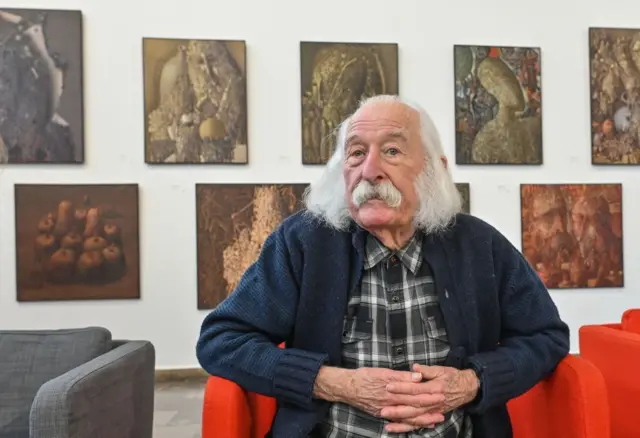 Іван Марчук у картатій сорочці та синьому светрі сидить на червоному дивані 