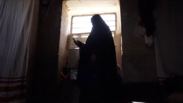 مژگان که چادر نماز بزرگ سیاه به سر دارد، مقابل پنجره ایستاده است. نور بیرون به داخل خانه میتابد و مژگان در حالی که تلفن به دست دارد، شماره میگیرد.
