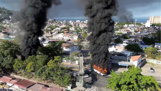 Fumaça sobe de ônibus em chamas no México. A imagem mostra a cidade ao longe, com o mar e o céu azul ao fundo