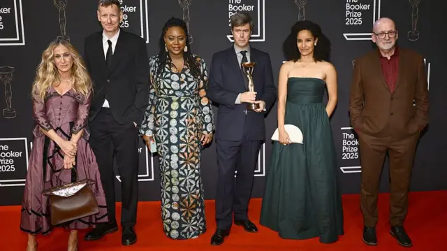 Лауреат Букеровской премии 2025 года Дэвид Солой, автор романа «Плоть» (в центре), позирует с членами жюри (слева направо): Сарой Джессикой Паркер, Крисом Пауэром, Айобами Адебайо, Кайли Рид и председателем жюри Родди Дойлом во время церемонии вручения Букеровской премии 2025 года 