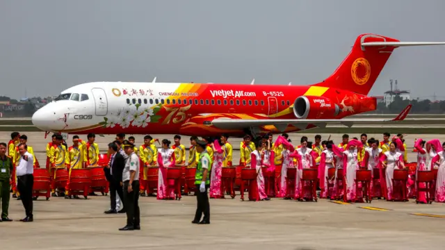 一架同时印上越捷航空标志的成都航空中国商飞C909客机在河内内排国际机场停机坪上，前面有身穿民族服装的越南人准备打鼓（14/4/2025）