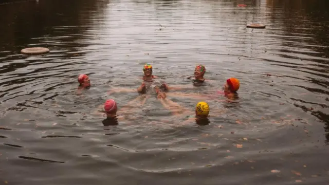 گروهی از شناگران در برکه‌ آب سرد با برگ‌های پاییزی در آب