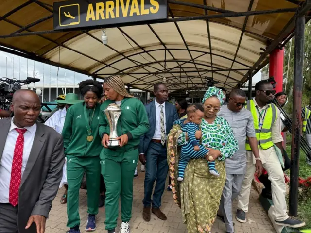 Foto of baby Bentley Taylor, Sarah Ogoke -Taylor pikin wey wear strip navy blue and light blue cloth dey for one woman wey carry am body while im mother and D'Tigress captain dey for back. Oda pipo mostly men dey di foto.