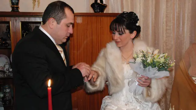 Жених в черном костюме надевает обручальное кольцо на палец своей новой жены. Она одела белое платье и белый меховой кардиган и держит букет белых лилий.