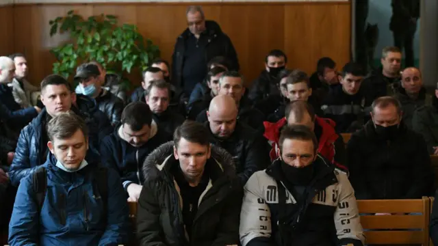 У чотири ряди сидять чоловіки різного віку з опущеними головами. Вони всі в цивільному.