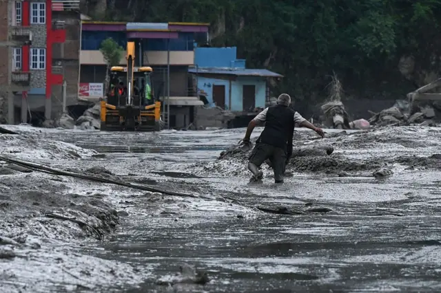 A person struggles through thick, grey mud and shallow floodwater in a heavily damaged urban area. In the background, a yellow excavator stands on the mud-covered route.