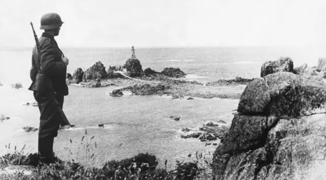 A German soldier stands on a rocky ledge, with a small rocky peninsula topped by a lighthouse in the background.