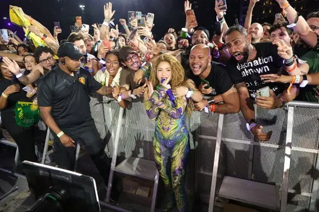Shakira cercada por fãs perto do palco no Rio de Janeiro. Ela usa uma roupa nas cores do Brasil e os fãs gritam e gesticulam. 