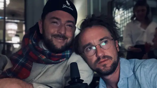 David w tartanowym szaliku i czapce Yankees obok Toma, który ma na sobie niebieską koszulę i okulary. Są w środku, w słabo oświetlonym pomieszczeniu.