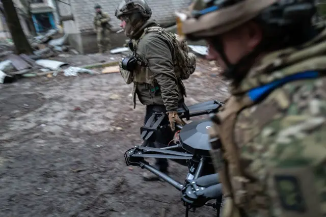 Двоє військових у камуфляжному спорядженні несуть великий багатокоптерний дрон з важкою рамою. Вони рухаються по сирому, вкритому багнюкою подвір’ю серед зруйнованих будівель та розкиданих уламків. На задньому плані видно ще одного військового біля стіни.