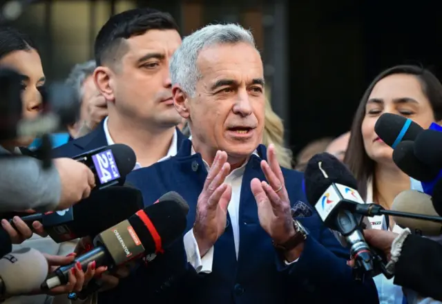 Muškarac u plavom odelu gestikulira rukama pred mikrofonima