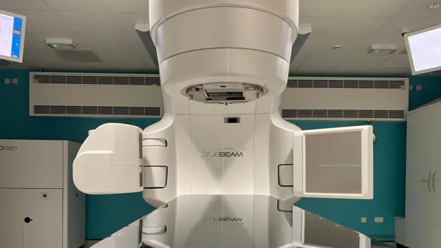 Una gran máquina de radioterapia color crema se encuentra en una habitación. Tiene una mesa plana y brillante para recostarse, con una máquina encima. En el techo hay dos pantallas de computadora. El logotipo dice "TrueBeam".