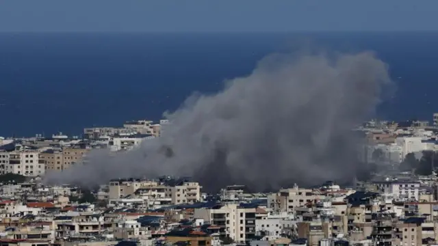 青い海と空を背に、建物が密集する市街地で巨大な灰色の煙が上っている