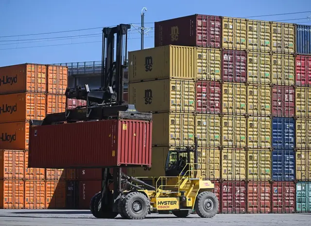 Una grúa transportando un contenedor en un puerto lleno de contenedores de carga.