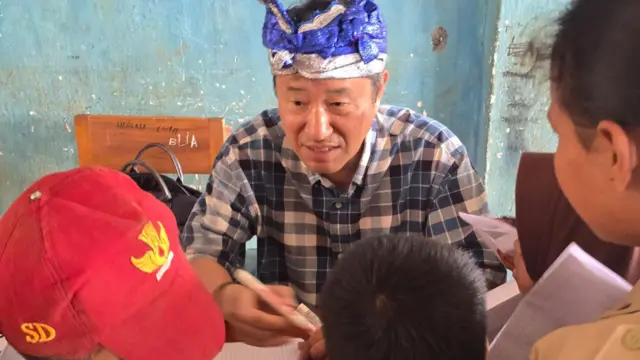 Jung Deuk Young, yang mengenakan kemeja kotak-kotak dan ikat kepala, duduk di hadapan tiga siswa sekolah dasar di Buton Selatan.