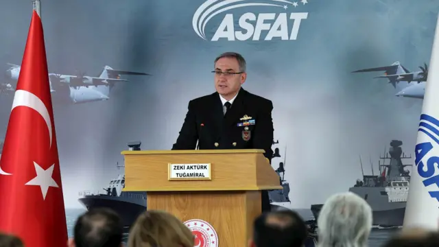 Tuğamiral Zeki Aktürk bir kürsüde konuşma yapıyor. Basın toplantısında kürsünün önünde arkasını dönük oturan katılımcılar var. 