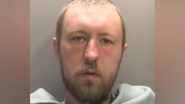 A mug shot of John Ashby who has short light brown hair and facial hair. he has a grey blanket across his front