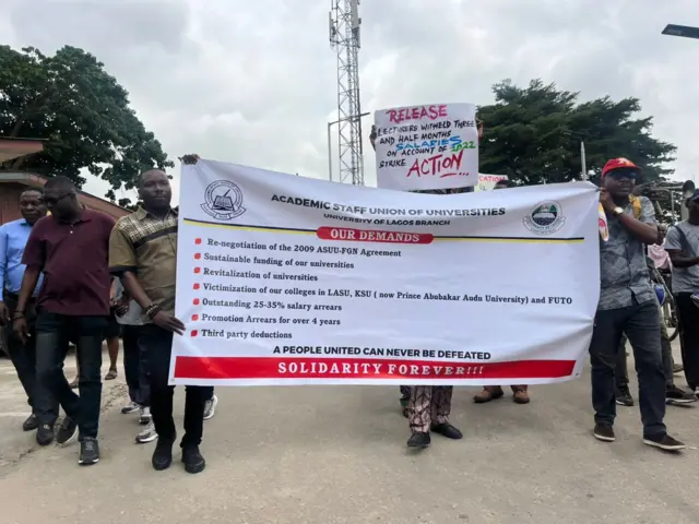 Members of di Academic Staff Union of Universities carry banner wey list all di tins dem want from goment.
