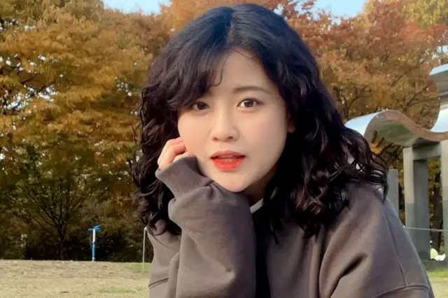 Baek Se-hee has shoulder-length curly dark hair and is sitting outdoors on grass, wearing a loose, long-sleeved gray sweatshirt. The background features autumn-coloured trees with shades of orange and yellow, and a metallic curved structure partially visible on the right side. She appears to be in a park or open recreational area during autumn.