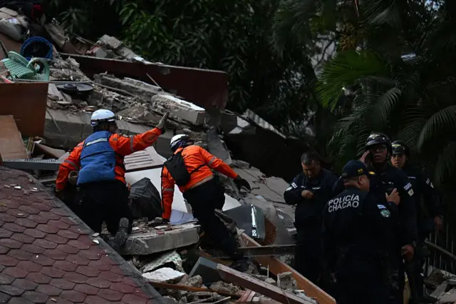 Equipos de rescate buscan entre los escombros de una estructura colapsada mientras agentes de la policía municipal permanecen cerca.