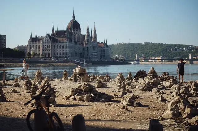 Una bicicleta y una persona en la parte del lecho del Danubio que la sequía ha hecho visible a su paso por Budapest. Más lejos, se aprecia el agua del río y un edificio histórico con una cúpula y varios pináculos bajo un cielo soleado.