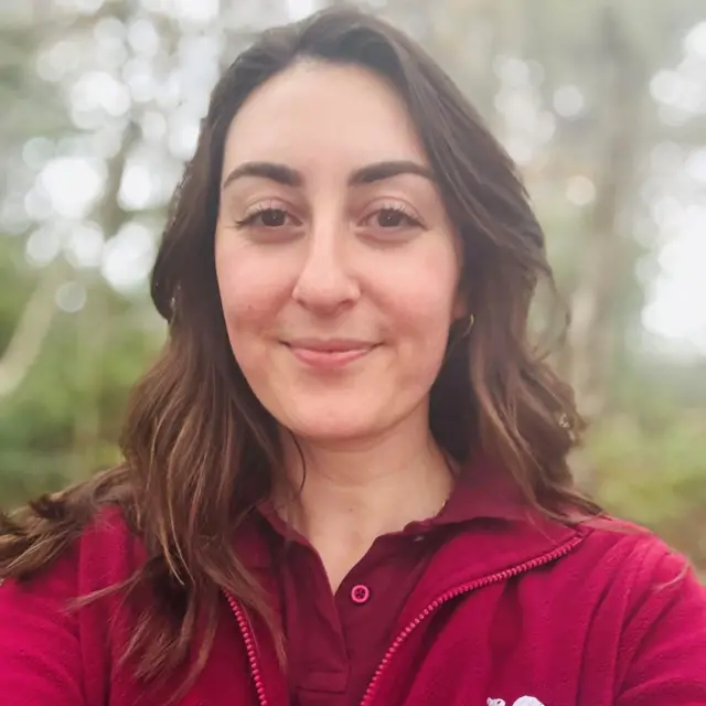 Naomi Bentley, de Saving Scotland's Red Squirrels, aparece fotografiada con una camiseta con cremallera de color granate brillante que tiene escrito 'Saving Scotland's Red Squirrels', sonriendo a la cámara, en el bosque.