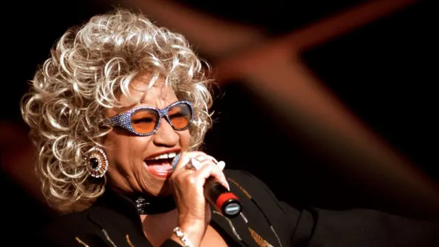 Celia Cruz, rubia, con unos lentes de sol cantando con un micrófono en la mano