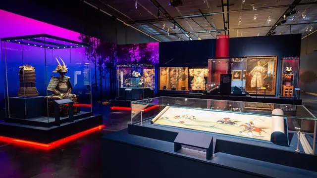 Parte de la muestra de la exposición “Samuráis” en el Museo Británico. En la imagen se aprecia, en primer plano y detrás de una vitrina, un documento ilustrado con figuras de personas cabalgando. A la izquierda se observa una armadura, y al fondo se distinguen cuadros y otras imágenes, aunque no con claridad.