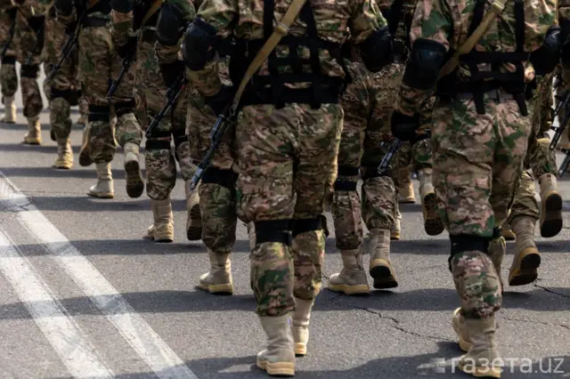 Rasmda saf tortib ketayotgan harbiy jihozlangan askarlarning orqa tomonidan tushirilgan tasviri