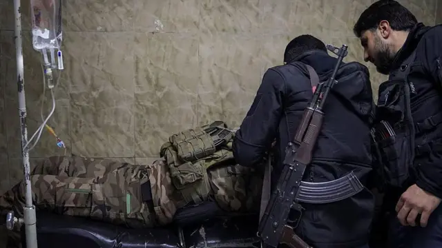 Suriye ordusuna bağlı bir asker, 6 Ocak 2026'da Halep'te yaralandıktan sonra hastaneye getirilmiş, yanında arkası dönük iki erkek asker var ikisi de silahlı ve siyah giyinmiş. Askeri üniformalı kişi sedyede uzanmış baş tarafı görünmüyor. Sol tarafta bağlı olduğu serum düzeneği var. 