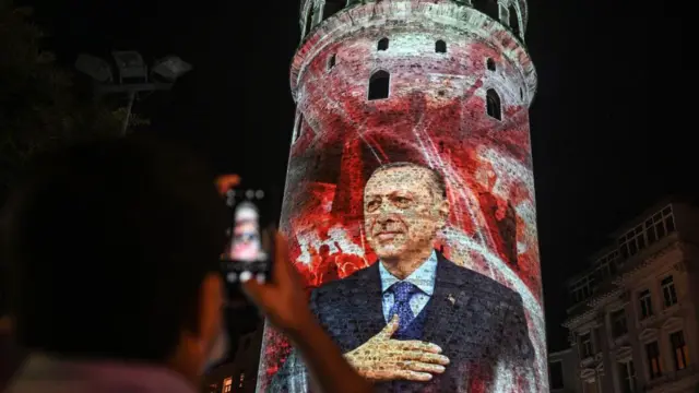 15 Temmuz 2021'de, İstanbul'daki Demokrasi ve Milli Birlik Günü kutlamaları kapsamında Galata Kulesi'ni gösteren, akşam çekilmiş bir fotoğraf. Kulenin üzerine Cumhurbaşkanı Recep Tayyip Erdoğan görüntüsü yansıtılmış. Bir kişi de gösteriyi cep telefonuyla kaydediyor.