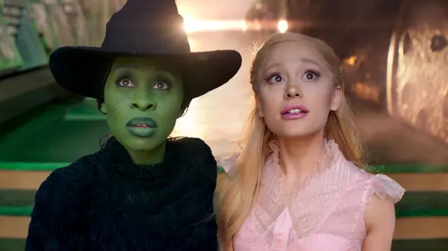 Cynthia Erivo e Ariana Grande olham para a câmera vestidas como suas personagens de Wicked, Elphaba e Glinda, em uma cena do filme Wicked. Elphaba usa uma roupa preta e um chapéu de bruxa preto, enquanto Glinda usa um vestido rosa