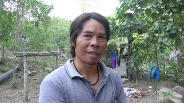 Junaidi berdiri di pekarangan dekat tanaman-tanaman. Dia mengenakan kaos polo berwarna abu-abu dan kalung manik berwarna hitam.