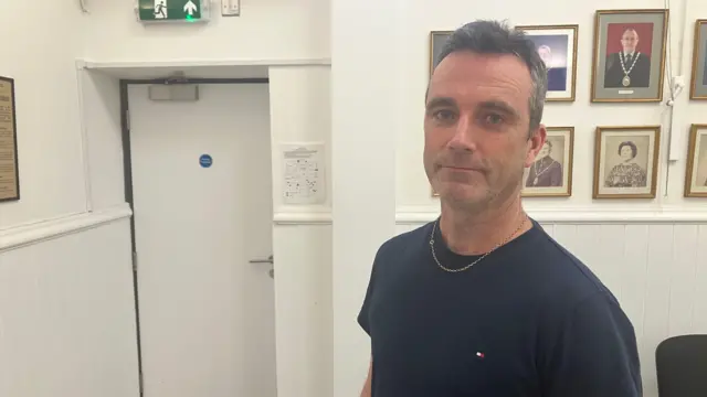 Un hombre con una camiseta azul marino se encuentra en un pasillo decorado con retratos enmarcados en la pared. A la izquierda hay una puerta blanca bajo una señal verde de salida de emergencia, y a la derecha hay varias sillas junto a la pared.